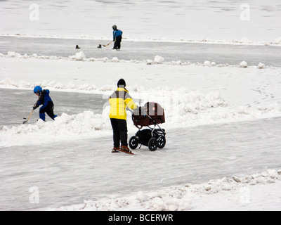 Woman with skates pushing a perambulator on a lake in Sweden. Kids playing hockey in the background. Stock Photo