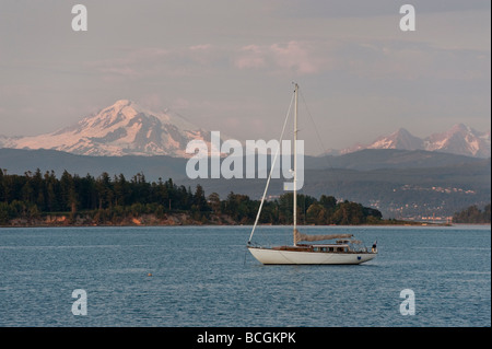 A sailboat anchors off Lummi Island, Washington. Snow capped Mt. Baker and the Lummi Indian Reservation dominate the background. Stock Photo