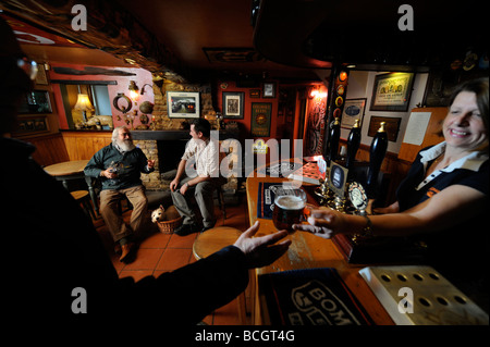 TWO MEN SIT BY AN OPEN FIRE IN A TRADITIONAL PUB AS THE LANDLADY HANDS A PINT OF ALE OVER THE BAR GLOUCESTERSHIRE ENGLAND UK Stock Photo