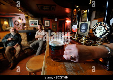 TWO MEN SIT BY AN OPEN FIRE IN A TRADITIONAL PUB AS THE LANDLADY PLACES A PINT OF ALE ON THE BAR GLOUCESTERSHIRE ENGLAND UK Stock Photo