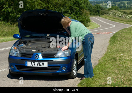 Lone woman looking under the bonnet of a broken down car at the side of the road Stock Photo