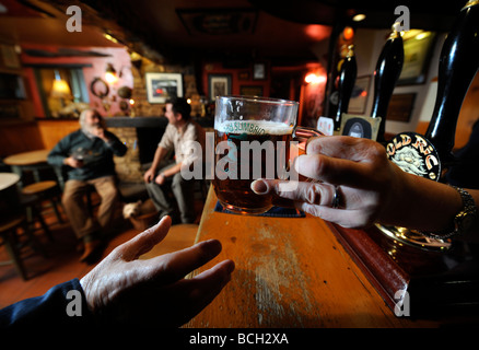 TWO MEN SIT BY AN OPEN FIRE IN A TRADITIONAL PUB AS THE LANDLADY SERVES A PINT OF ALE OVER THE BAR GLOUCESTERSHIRE ENGLAND UK Stock Photo