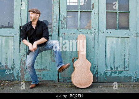 A musician does a funny dance if front of a green building.  His guitar is leaning against the building in a brown case. Stock Photo