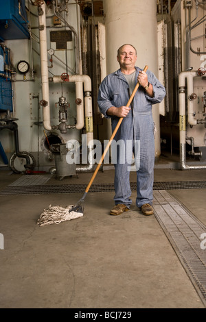 Male Caucasian in blue custodian/janitor overalls in factory setting including boiler, pipes, valves.  He is holding mop. Stock Photo