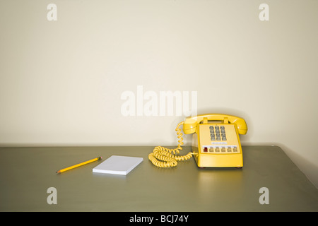 Yellow touch tone telephone sitting on metal desk in front of white wall.  Pencil and pad of paper sitting next to phone on desk Stock Photo