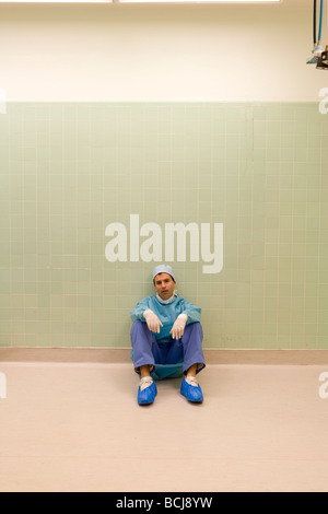 Caucasian male surgeon/doctor in blue surgical scrubs sitting on floor against green tile wall, tired, exhausted. Stock Photo