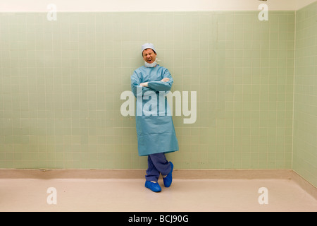 Caucasian male surgeon/doctor in blue surgical scrubs leaning against green tile wall yawning. Stock Photo