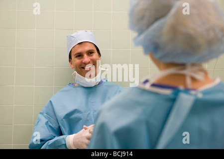 Caucasian male surgeon/doctor in blue surgical scrubs leaning against green tile walk, speaking with second person. Stock Photo