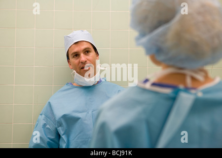 Caucasian male surgeon/doctor in blue surgical scrubs leaning against green tile walk, speaking with second person. Stock Photo