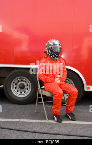 Race car driver wearing helmet and red jump suit sitting in chair in front of red transport vehicle  Stock Photo