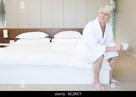 Middle aged man in bathrobe sitting on bed with cup of coffee Stock Photo