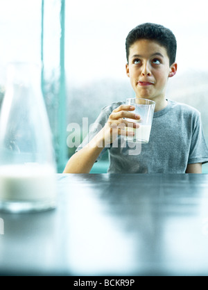 Boy sitting at table holding glass of milk and licking lips, carafe of milk in foreground Stock Photo
