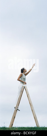 Woman on ladder, reaching toward sky Stock Photo