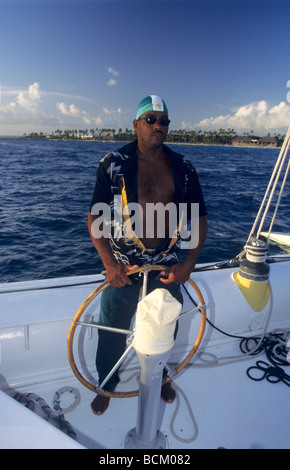 Black sailor man steering at the helm on a catamaran cruise ship - Dominican Republic Island -  Bayahibe coast - Caribbean sea Stock Photo