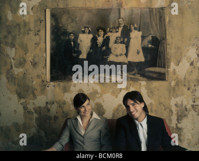 Man and woman sitting side by side, smiling, under black and white family photo on wall Stock Photo