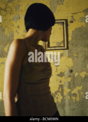 Woman looking over shoulder at frame on flaking wall, focus on background Stock Photo