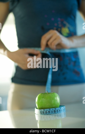 Woman standing with one end of measuring tape around waist and other end around apple on counter in front of her, cropped view Stock Photo