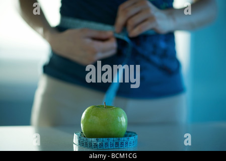 Woman standing with one end of measuring tape around waist and other end around apple on counter in front of her, cropped view Stock Photo