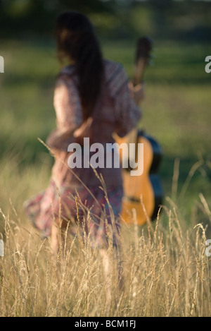 Rear view of woman walking through field, carrying guitar, focus on tall grass in foreground