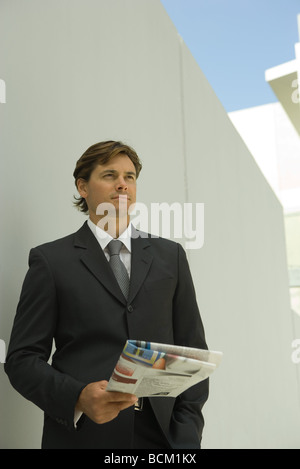 Businessman standing outdoors, holding newspaper, looking away Stock Photo