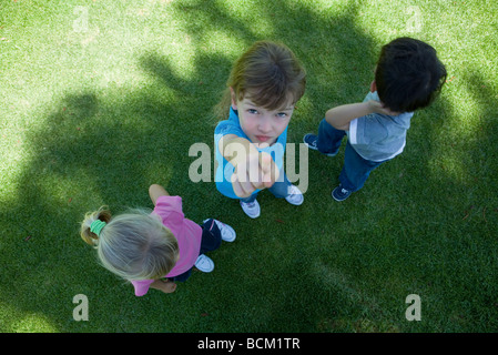 Three children standing on grass, in shade, one girl pointing at camera, view from directly above Stock Photo