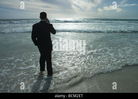 Businessman standing in surf at beach, using cell phone, rear view Stock Photo