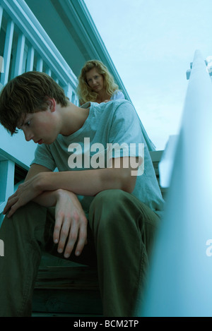 Teenage boy sulking on steps, head down, mother standing in background Stock Photo