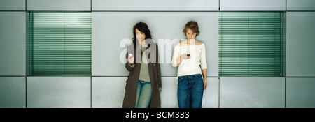 Two young females leaning against wall, both text messaging with cell phones Stock Photo