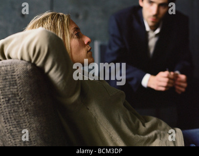 Young woman reclining on couch, psychologist looking at her in background Stock Photo