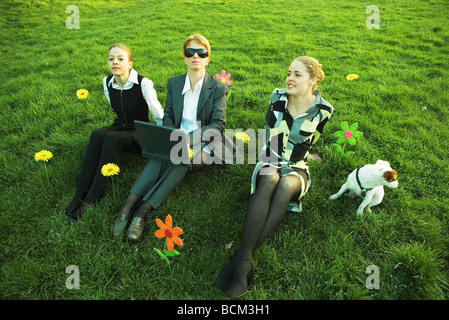 Three females sitting side by side with dog in field with flowers, one using laptop computer, high angle view Stock Photo