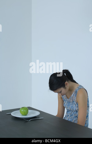 Woman sitting at table with head down, single apple on plate before her Stock Photo