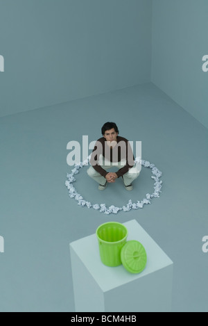 Man crouching inside circle of paper balls, garbage can in foreground, high angle view Stock Photo