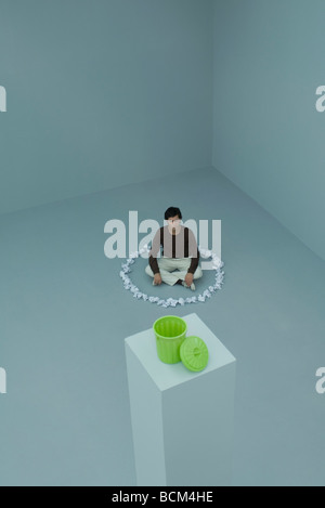 Man sitting on the ground inside circle of crumpled paper balls, garbage can on pedestal in foreground Stock Photo