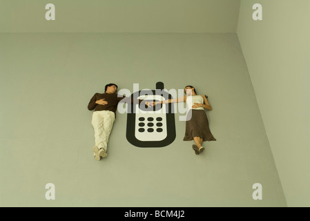 Couple lying on the ground with large cell phone between them, holding hands Stock Photo