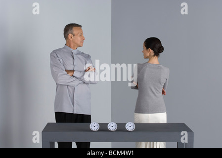 Couple standing with arms folded, looking at each other, clocks on table in front of them Stock Photo