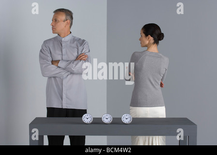 Couple standing with arms folded, backs turned to each other, clocks on table in front of them Stock Photo