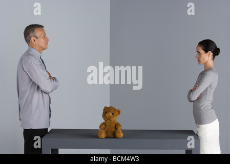 Couple standing face to face at table, arms folded, teddy bear between them Stock Photo