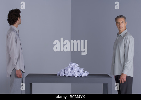 Two men standing at opposite ends of table, pile of crumpled paper between them, both looking away Stock Photo