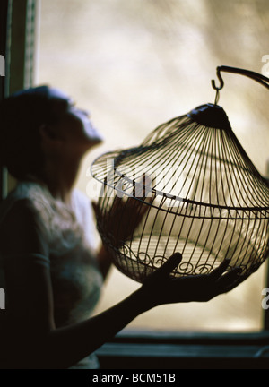 Woman standing beside window, holding empty birdcage Stock Photo