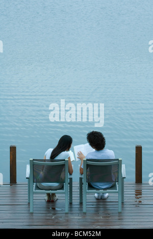 Couple sitting side by side on dock, reading books, rear view Stock Photo