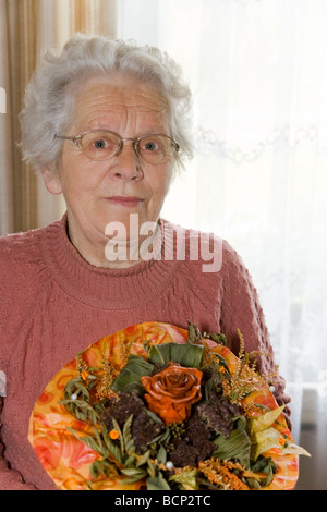 Frau in ihren Siebzigern steht im Wohnzimmer und hält einen künstlichen Blumenstrauß Stock Photo