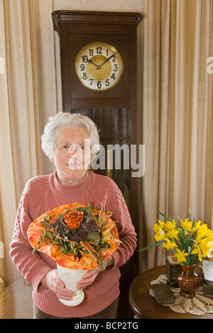 Frau in ihren Siebzigern steht im Wohnzimmer und hält einen künstlichen Blumenstrauß Stock Photo