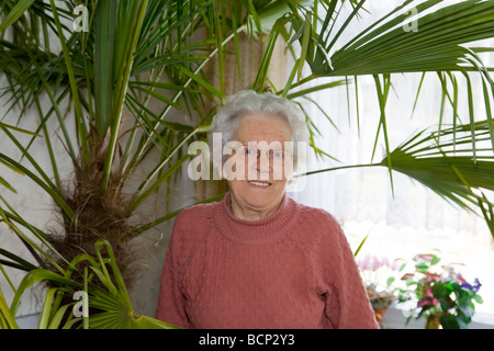 Frau in ihren Siebzigern steht vor einer großen Palme im Wohnzimmer Stock Photo
