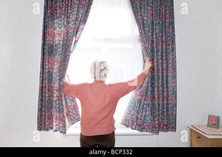 Frau in ihren Siebzigern steht morgens am Fenster und zieht die Vorhänge zur Seite Stock Photo