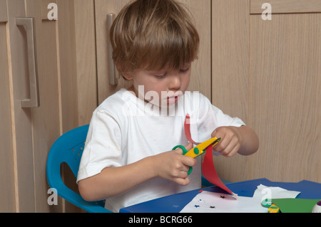 Three year old boy cutting with scissors sitting at small table in kitchen Stock Photo
