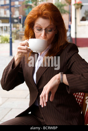 Busy professional businesswoman with red hair checking the time whilst drinking a cup of coffee Stock Photo