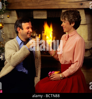 MR COUPLE CLINKING WITH CHAMPAGNE GLASSES AND EXCHANGING CHRISTMAS PRESENT BY FIREPLACE Stock Photo