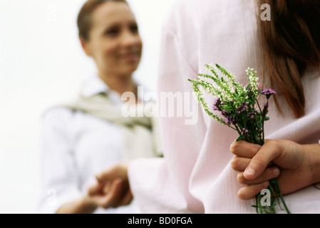 Girl hiding wildflower bouquet behind back, holding mother's hand, cropped