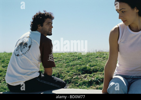 Young couple sitting apart on park bench, glancing over shoulders at one another Stock Photo
