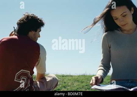 Young woman sitting outdoors reading magazine, man using cell phone behind her Stock Photo
